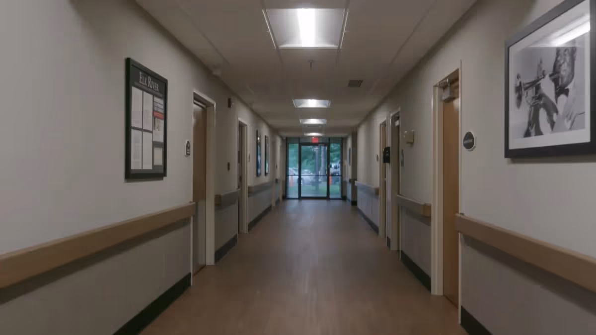 A long, well-lit hallway in a healthcare or nursing facility with wooden doors on both sides, handrails along the walls, a bulletin board on the left wall, and a framed black and white photo of a man playing a trumpet on the right wall. The hallway leads to glass exit doors with a view of greenery outside.