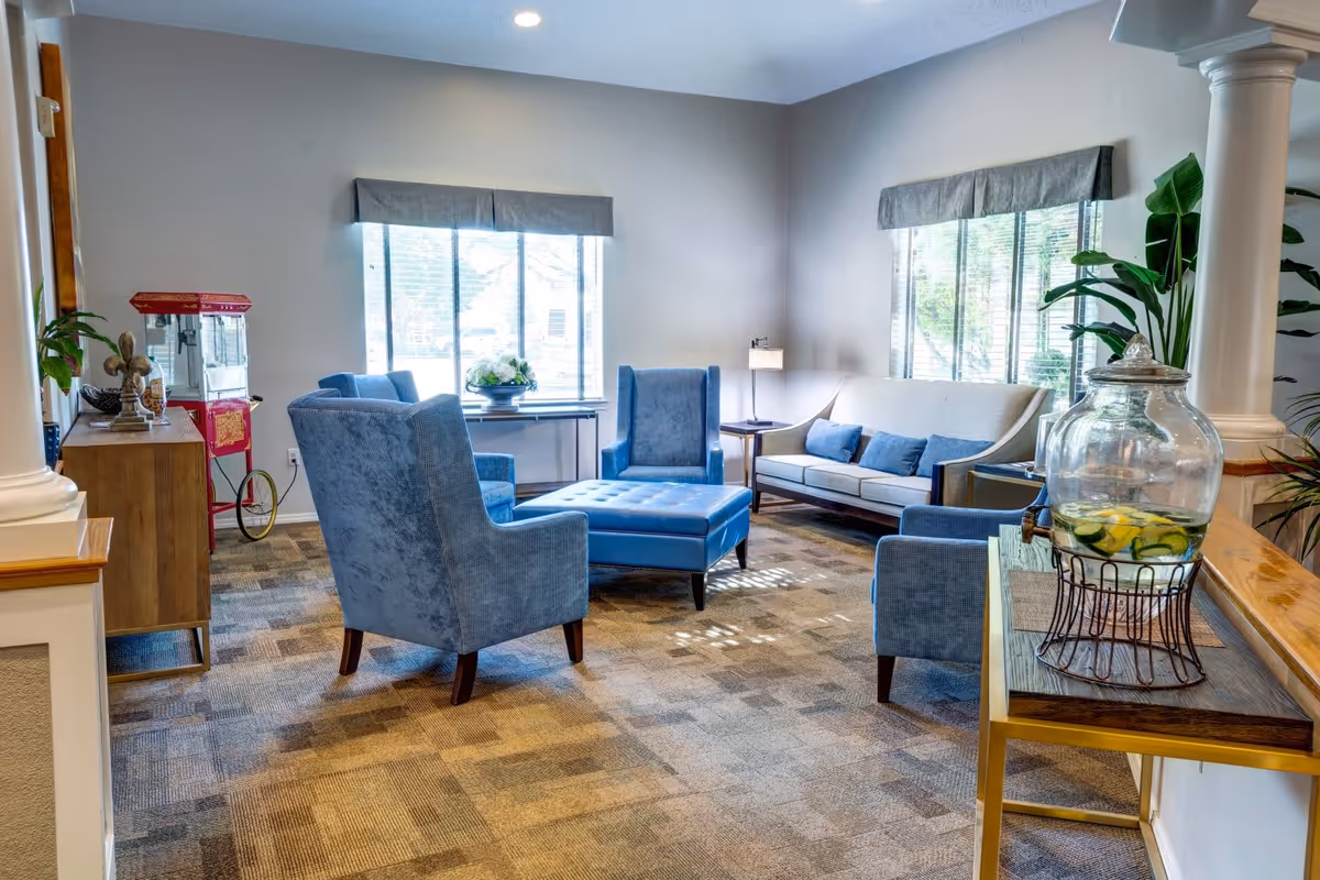 A bright and cozy living room area with blue upholstered chairs, a matching ottoman, a beige sofa with blue cushions, and large windows with gray valances letting in natural light. There is a wooden sideboard with decorative items and a red popcorn machine in the corner. A glass beverage dispenser with lemon and cucumber water sits on a wooden console table near white columns and green plants.