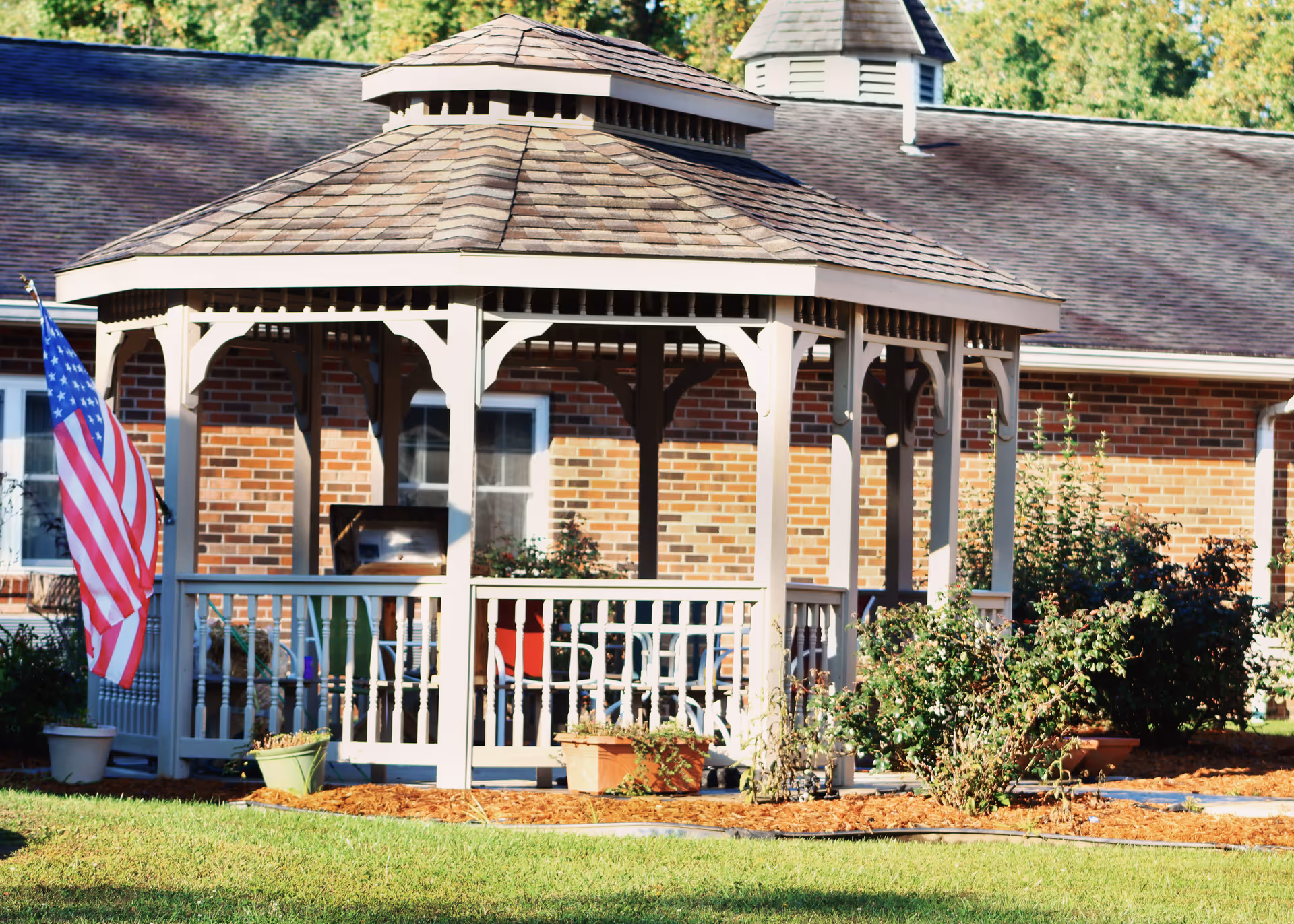 A beige wooden gazebo with a shingled roof situated in a garden area in front of a brick building. The gazebo has white railings and several potted plants around it. An American flag is displayed on the left side of the gazebo. Green grass and bushes surround the area.