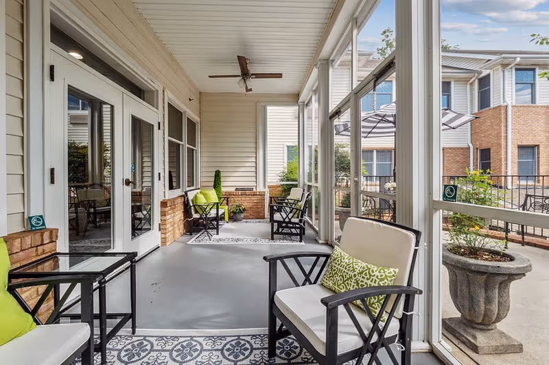 A screened-in porch area with cushioned chairs featuring green patterned pillows, a ceiling fan, and a small table. The porch overlooks an outdoor patio with umbrellas and seating, surrounded by residential-style buildings.