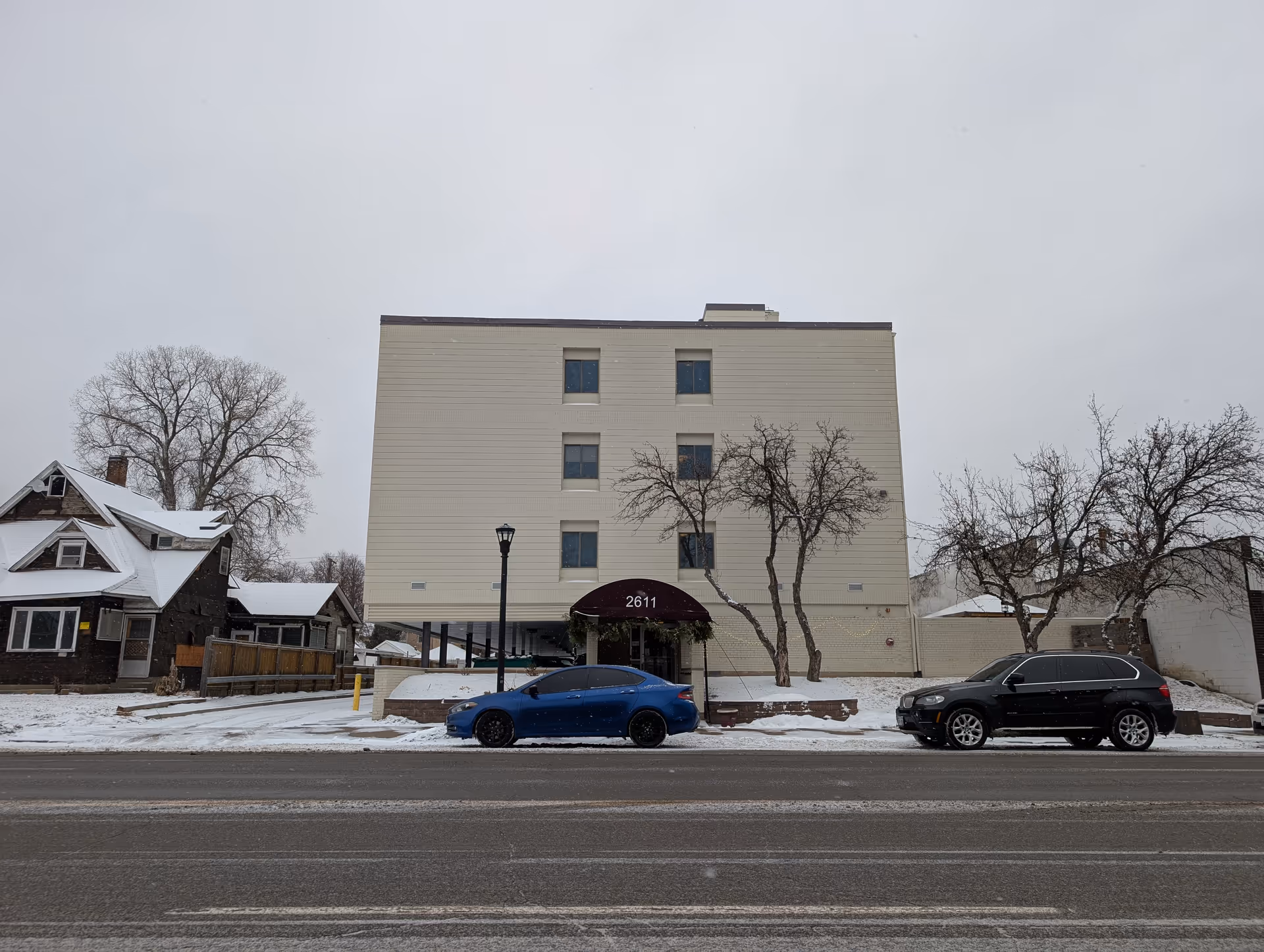 Four-story light-colored apartment building with an awning reading '2611', trees in front, and parked cars along a snowy street.