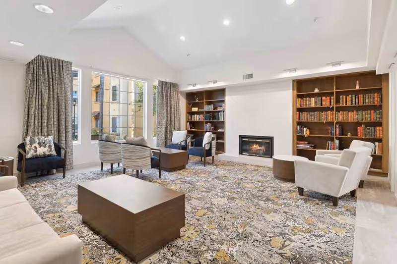 A spacious and well-lit living room with a high ceiling, large windows with curtains, a patterned carpet, multiple chairs and sofas arranged around a central wooden coffee table, and built-in wooden bookshelves flanking a modern fireplace.