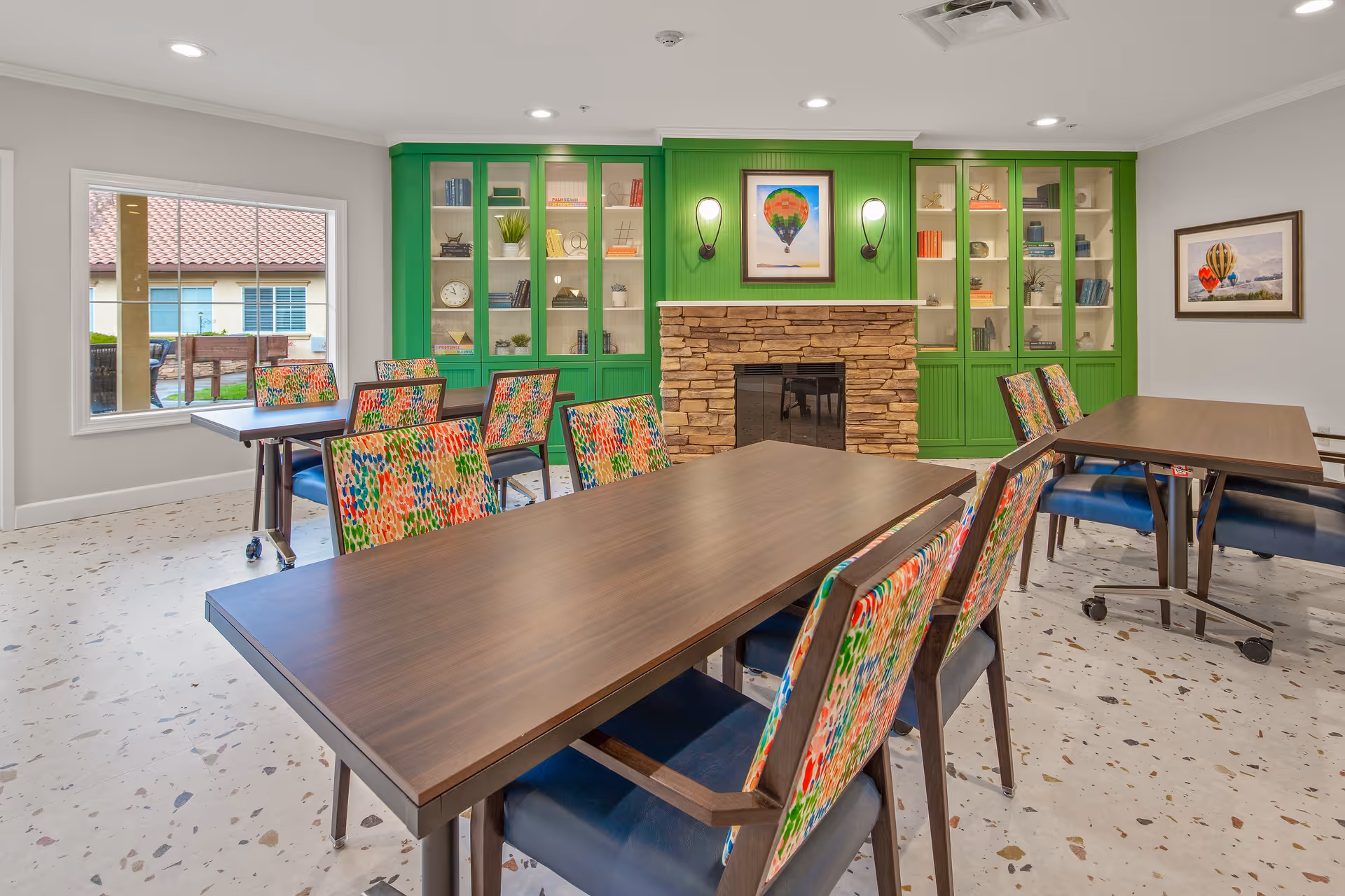 A bright and colorful common area with multiple wooden tables and chairs featuring multicolored patterned upholstery. The room has a stone fireplace centered on a green wall with built-in glass cabinets on either side displaying books and decorative items. There are two framed pictures of hot air balloons on the walls and a window showing an outdoor view.