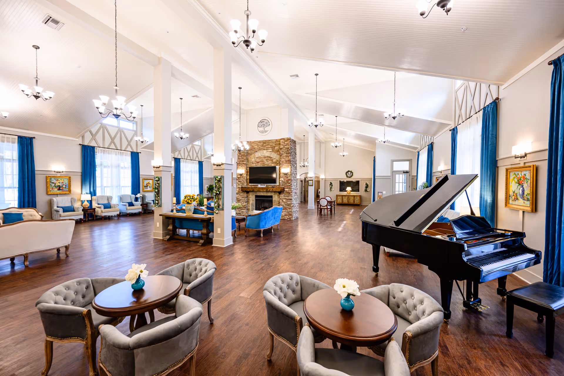 Spacious senior living common area with high vaulted ceilings, multiple seating arrangements including round tables with gray upholstered chairs, a grand piano, a stone fireplace with a mounted TV above it, large windows with blue curtains, and wooden flooring.