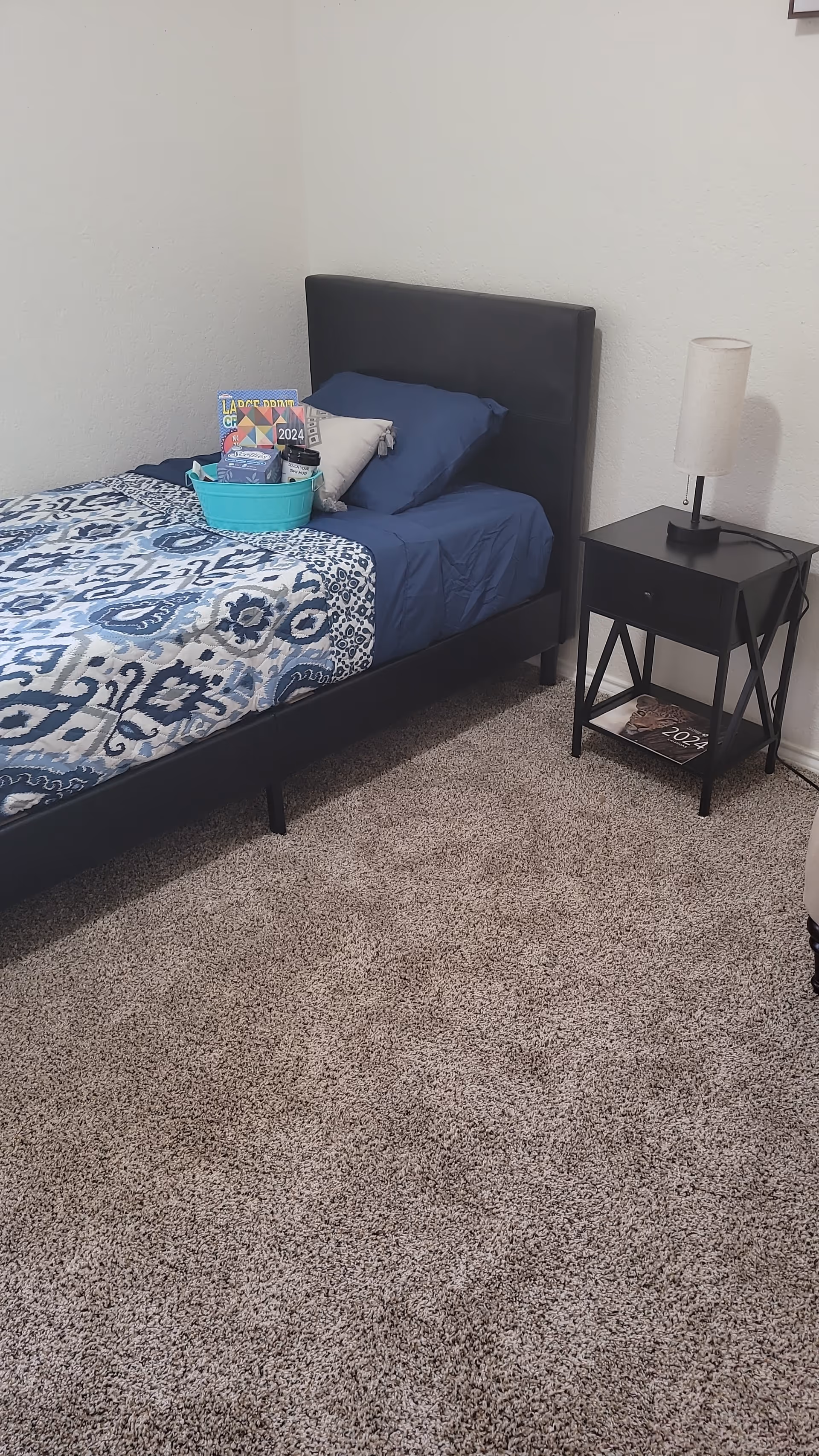 A small bedroom with a single bed dressed in blue patterned bedding, a nightstand with a lamp, and a gift basket on the bed.