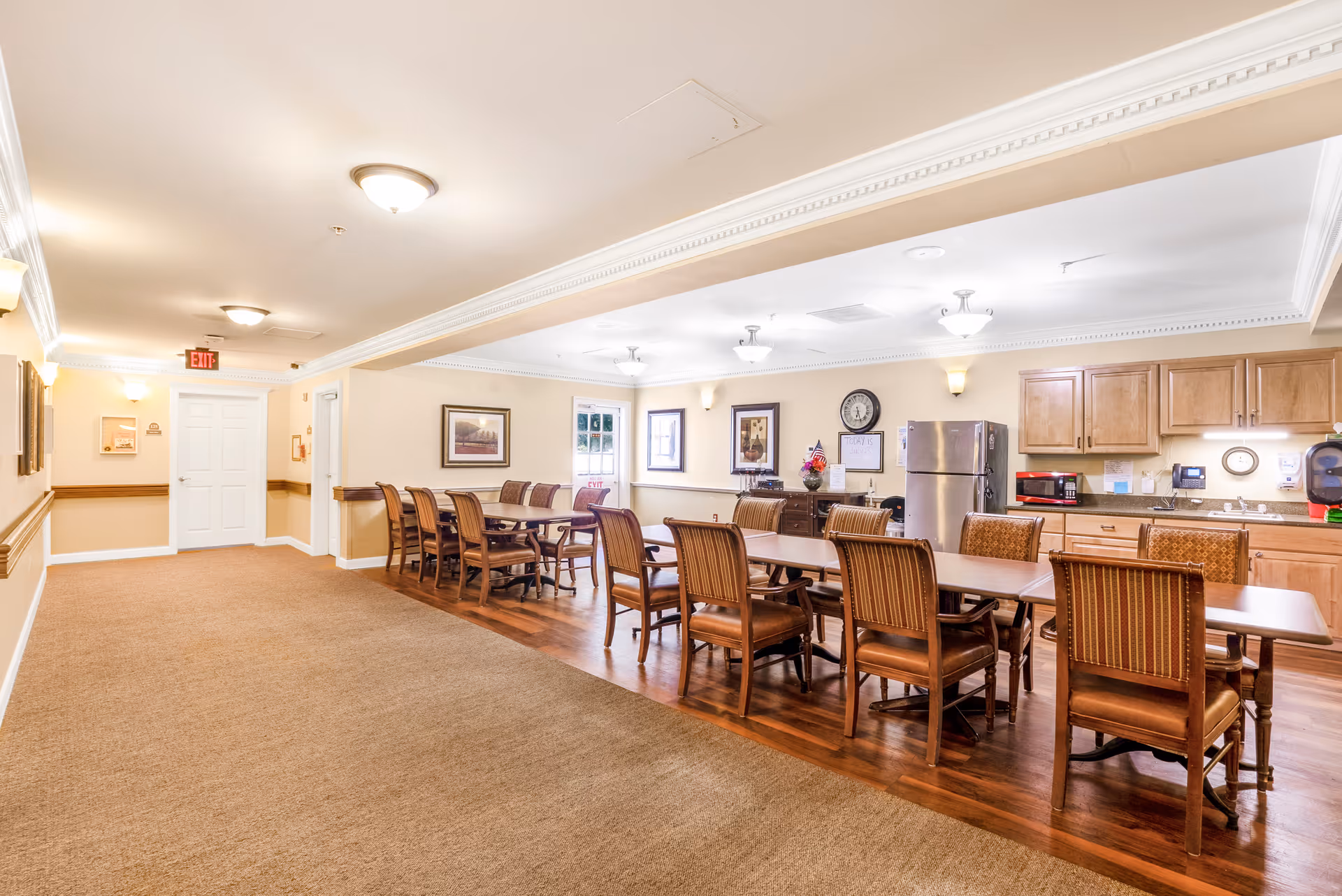 Communal dining area with long wooden tables and chairs beside a kitchenette and refrigerator.