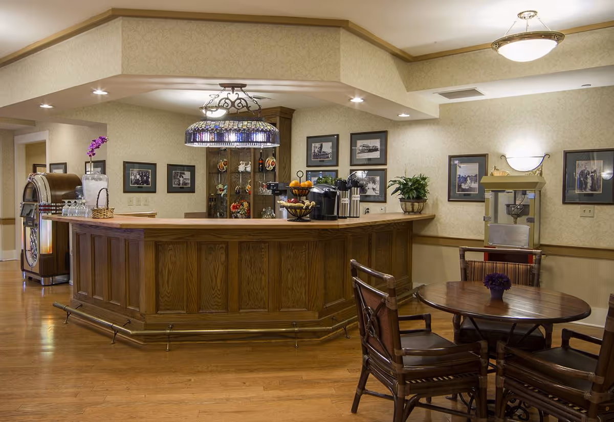 Interior view of a senior living facility lounge area featuring a wooden bar counter with coffee machines and fruit baskets on top. There is a round wooden table with four chairs in the foreground, a popcorn machine against the wall, framed pictures hanging on the wallpapered walls, and a vintage jukebox in the background.