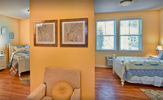 Interior view of a senior living facility bedroom area with two separate beds in adjacent rooms separated by a partial wall. Each room has wooden flooring, framed artwork on the walls, windows with blinds, and bedside tables with lamps. A beige armchair with a round pillow is positioned in front of the partial wall.