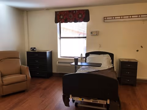 A simple room with a hospital-style bed, a brown recliner chair, two dark wooden dressers, a window with blinds and a decorative valance, and a wall-mounted air conditioning unit below the window. The floor is wooden, and the walls are light-colored.