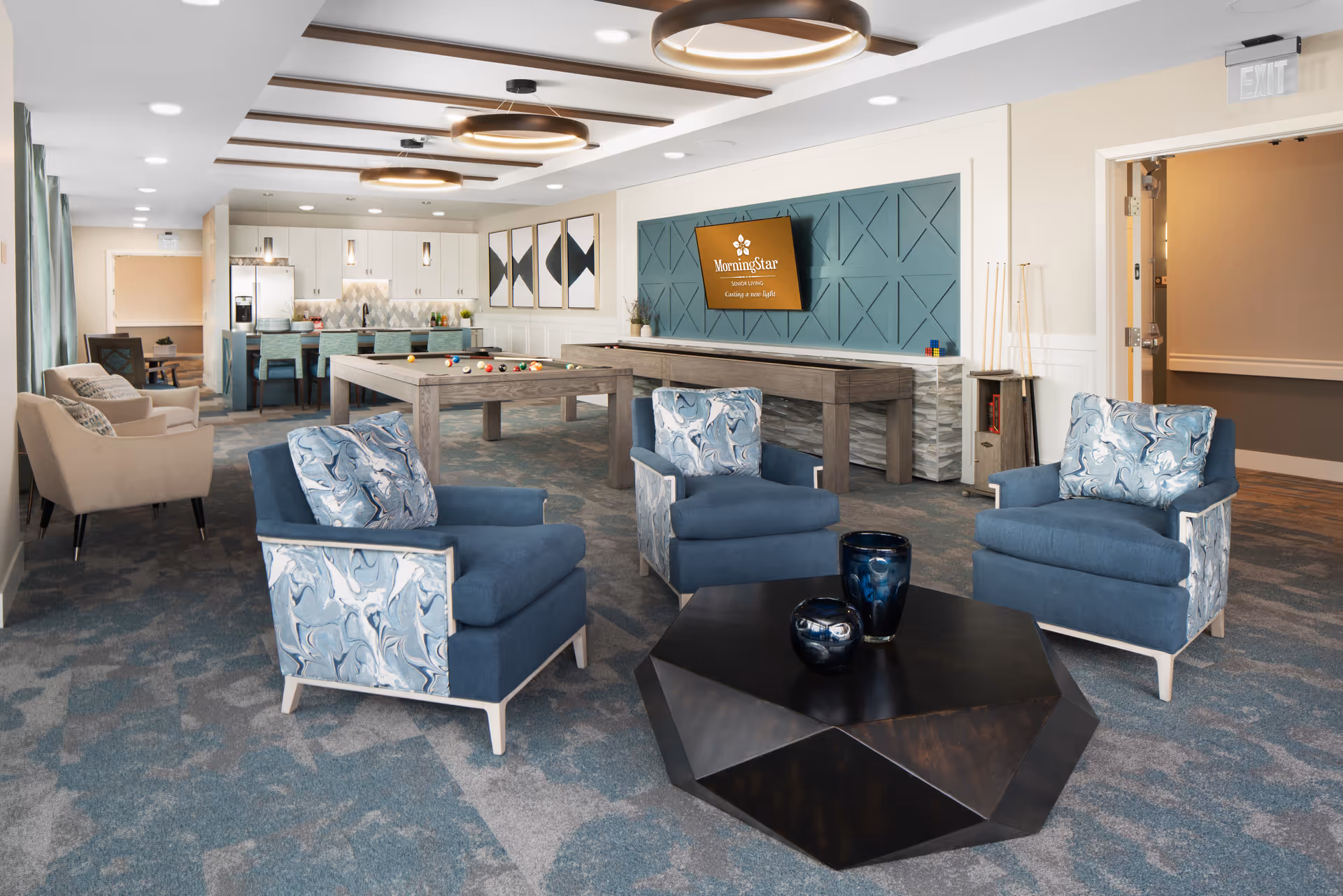 A spacious and modern common area in an assisted living facility featuring three blue armchairs with patterned sides around a geometric black coffee table with decorative vases. In the background, there is a pool table, a shuffleboard table, and a kitchen area with a counter and bar stools. The walls are decorated with a teal panel and a mounted screen displaying the MorningStar logo. The room has a carpeted floor and ceiling lights with wooden beams.
