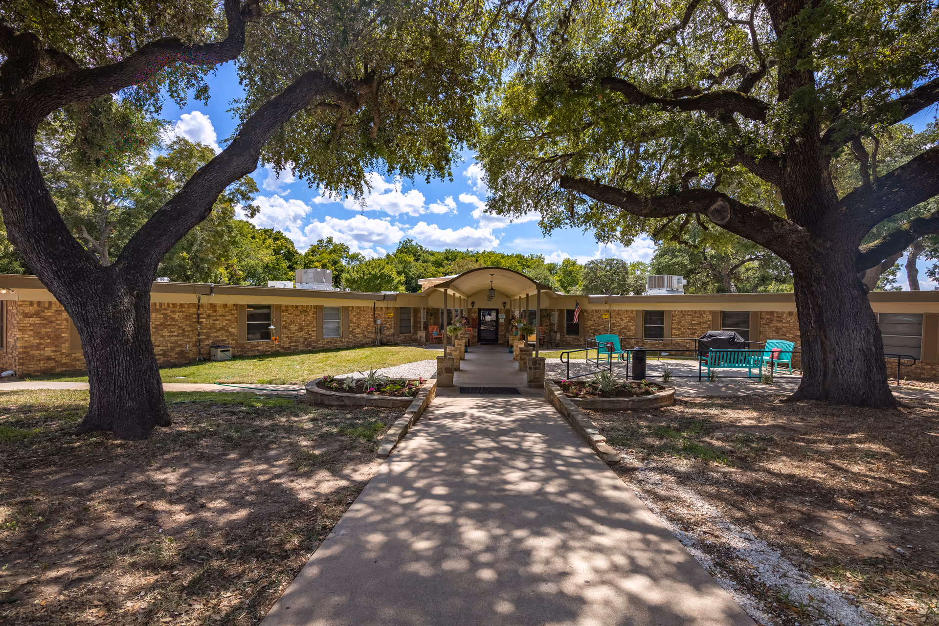 Exterior view of Care Inn of La Grange showing a single-story brick building with a covered entrance walkway. Large trees provide shade over a concrete path leading to the entrance. There are flower beds on either side of the walkway and outdoor seating with turquoise benches to the right.