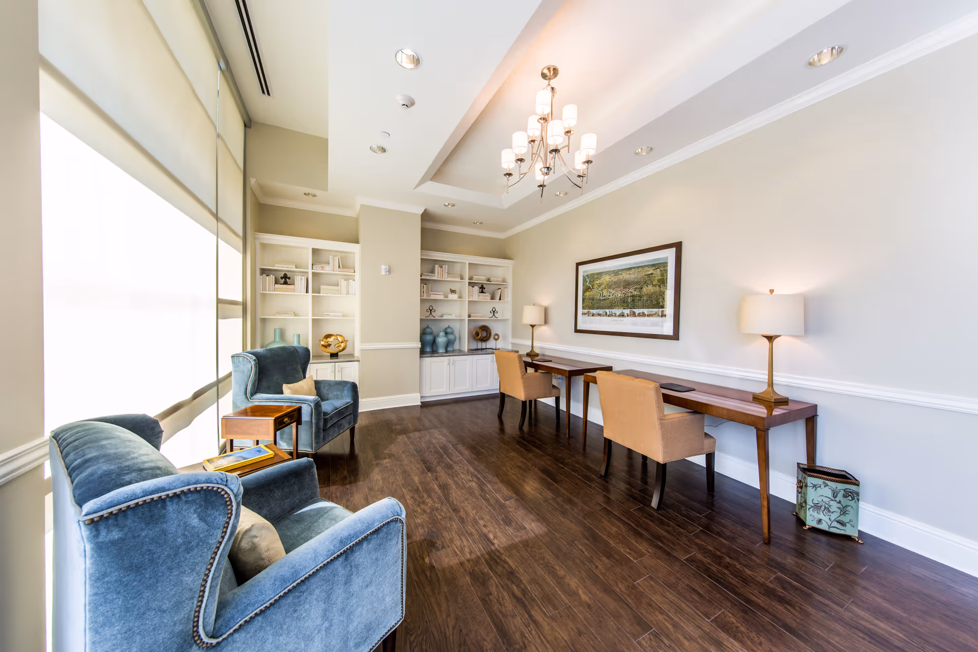 A bright and elegant interior room with two blue upholstered armchairs and a small wooden side table near large windows with blinds. On the opposite wall, there are two wooden desks with beige chairs and table lamps, a framed landscape painting above, and built-in white shelves with decorative items. The room has dark wooden flooring and a chandelier hanging from a recessed ceiling.