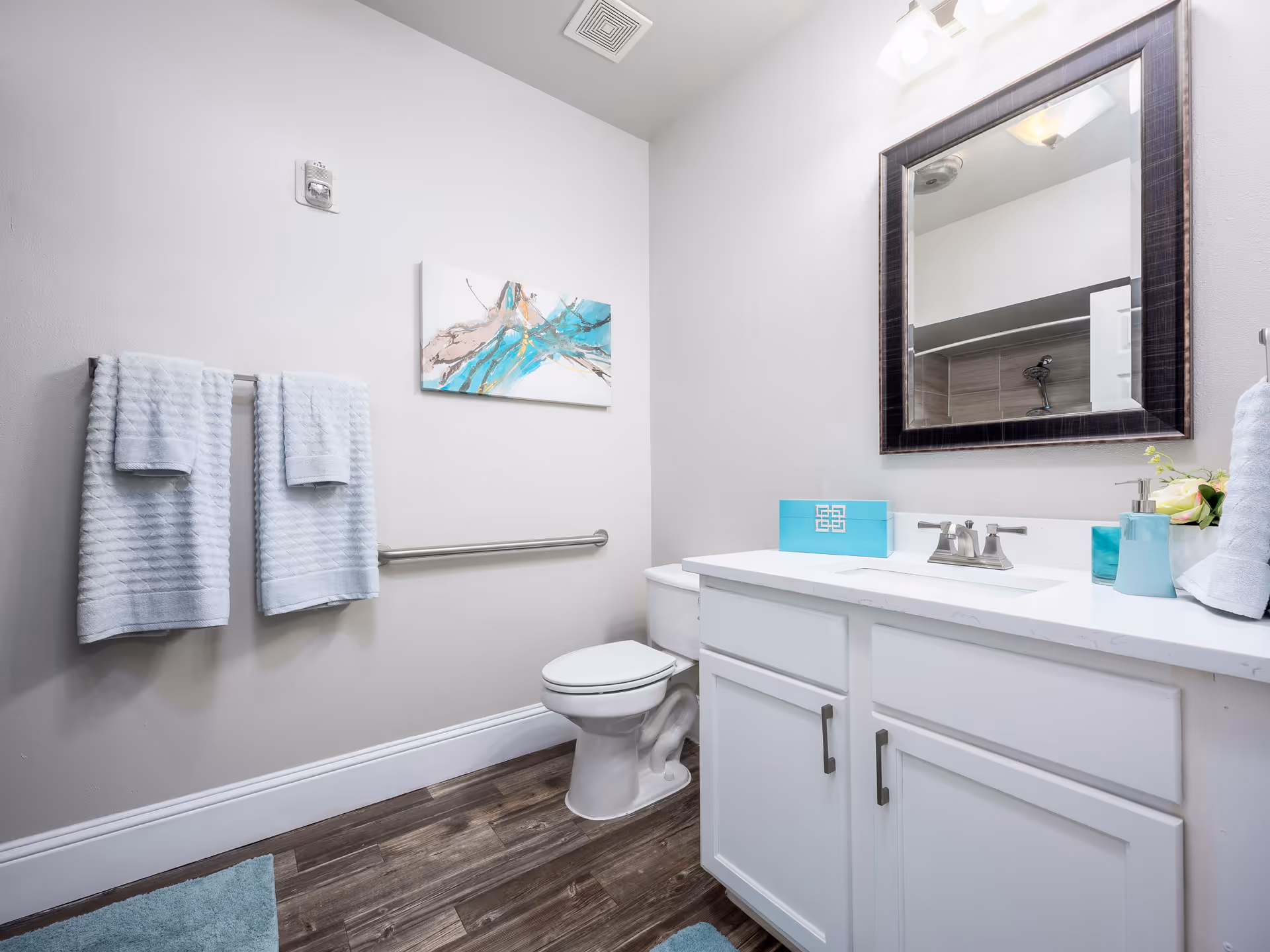 Bright modern bathroom with a white vanity, mirror, toilet, and teal-accented towels and accessories.