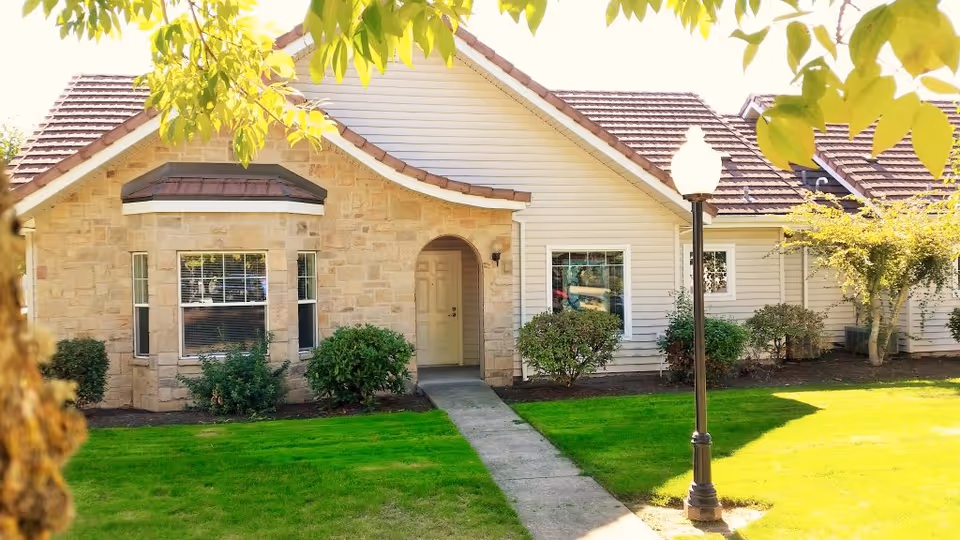 Exterior view of a single-story building with a stone and siding facade, a tiled roof, a front door under an arched entryway, surrounded by green grass, bushes, and trees with a lamppost near the walkway leading to the entrance.