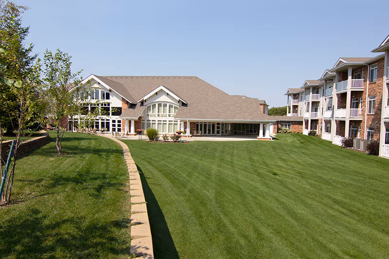 A wide, manicured lawn and paved path leading to a large multi-story senior living building and central clubhouse.