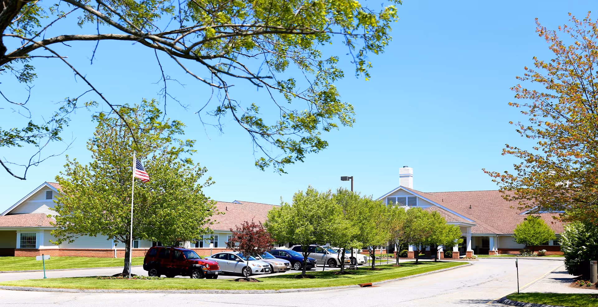Exterior view of a single-story building with a sloped roof, surrounded by green trees and a parking lot with several cars. An American flag is flying on a flagpole near the building under a clear blue sky.
