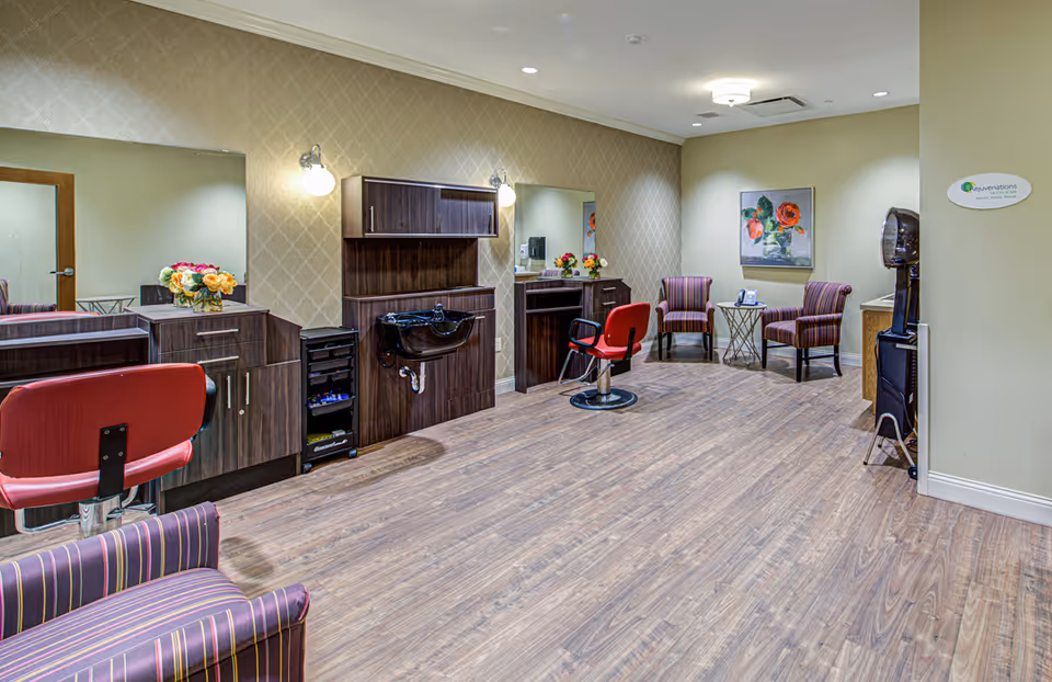 Interior view of a salon area in a senior living facility with two red salon chairs in front of large mirrors, dark wood cabinetry, and a black hair washing sink. There are two striped armchairs and a small round table with a phone in the corner, a floral painting on the wall, and a hair dryer station on the right side.