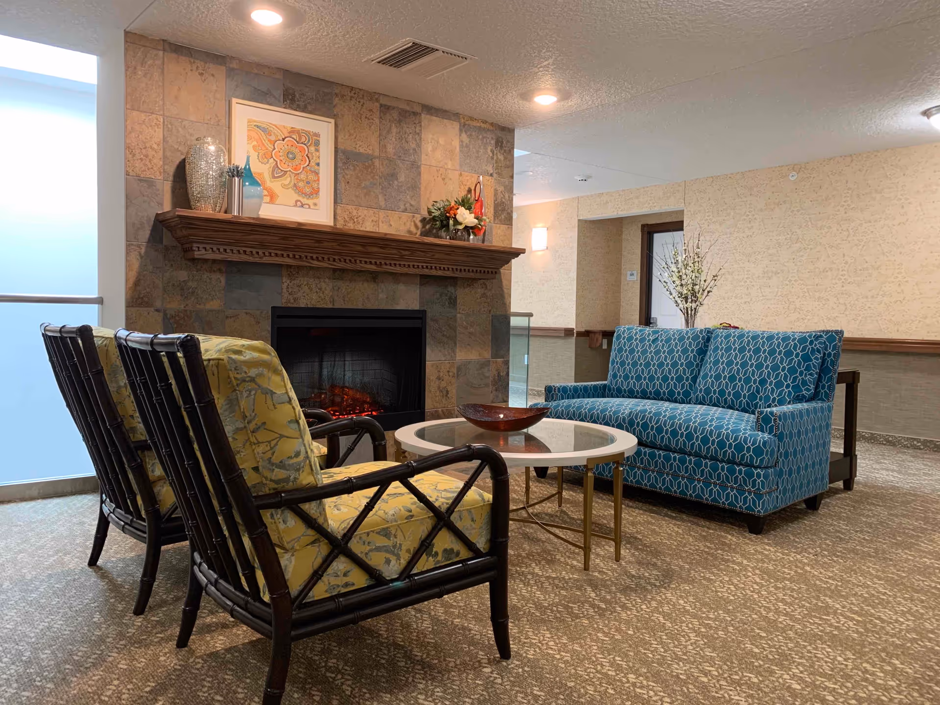 A cozy lounge with patterned armchairs and a blue loveseat around a round coffee table in front of a tiled fireplace.