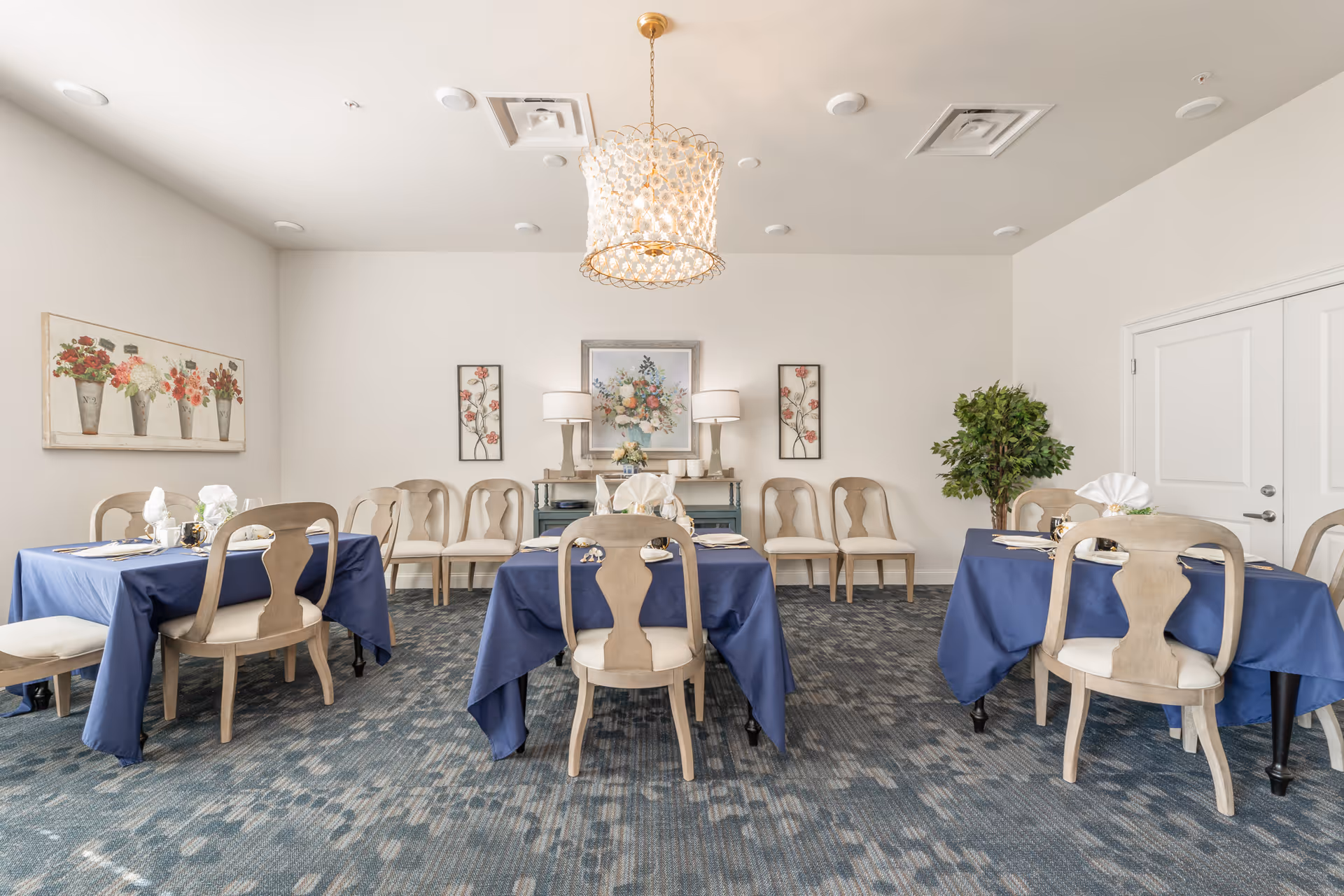 Bright dining room with tables covered in blue tablecloths, wooden chairs, floral artwork, a chandelier, and set place settings.