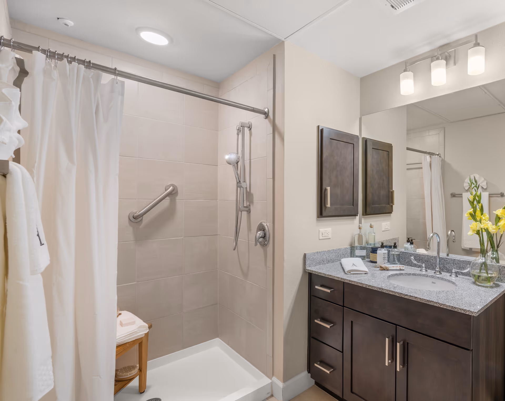 A clean and modern bathroom featuring a walk-in shower with beige tiles, a silver grab bar, and a handheld showerhead. There is a white shower curtain partially drawn, a wooden stool with folded towels, and a dark wood vanity with a granite countertop, sink, and a large mirror above. A vase with yellow and white flowers is placed on the countertop.