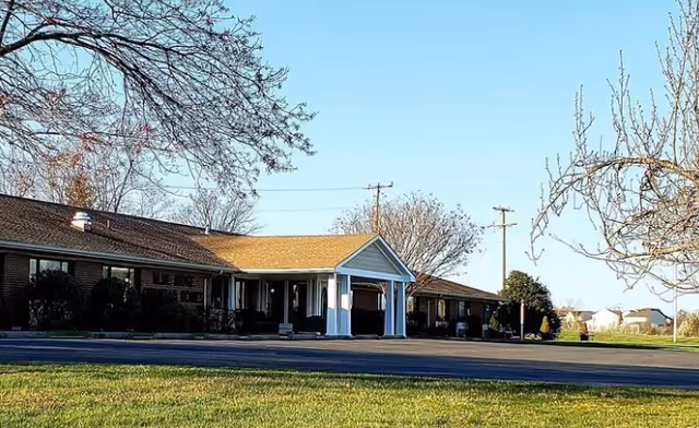Single-story brick senior living building with a covered entrance, lawn, and trees under a clear sky.