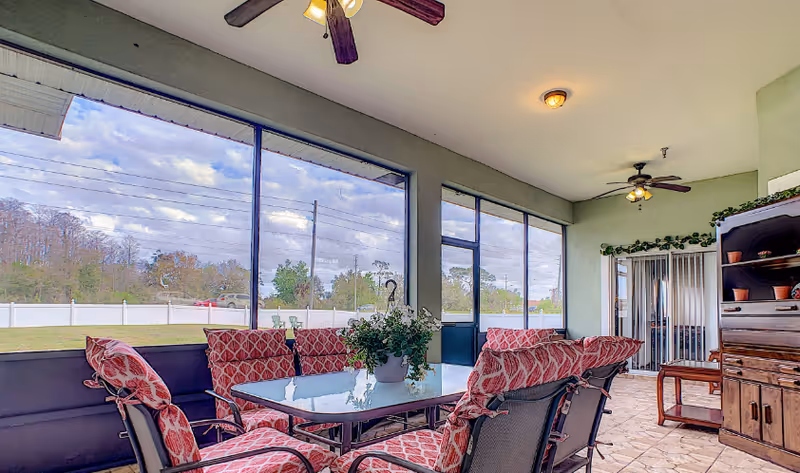 Sunroom-style covered patio with a glass-top dining table, patterned cushioned chairs, potted plant, ceiling fans, and large screened windows overlooking a yard.