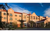 Exterior view of a multi-story senior living facility building with a red roof, beige walls, and decorative architectural details. The entrance features a covered drop-off area, surrounded by palm trees and landscaped greenery under a clear sky.