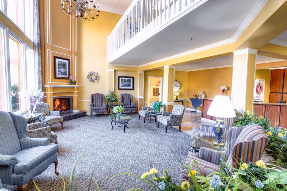 A spacious and well-lit living room area with high ceilings and large windows. The room features a lit fireplace with a framed picture above it, several upholstered armchairs arranged around glass coffee tables, and a chandelier hanging from the ceiling. There are plants and flowers decorating the space, and a reception desk is visible in the background.
