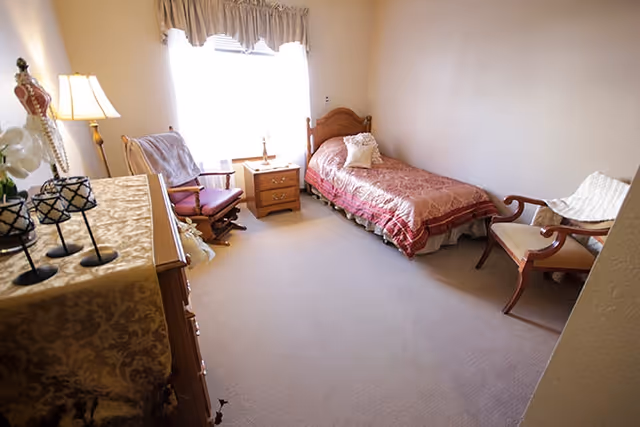 A small, cozy bedroom with a single bed covered in a red patterned bedspread, a wooden nightstand with a lamp, two wooden chairs with cushions, a dresser with decorative items, and a window with sheer curtains letting in natural light.