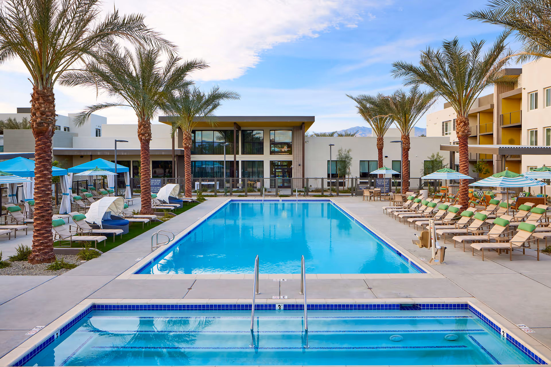 Outdoor swimming pool area at Revel Palm Desert with palm trees lining both sides, lounge chairs with green cushions, umbrellas, and cabanas. A modern building is visible in the background under a partly cloudy sky.