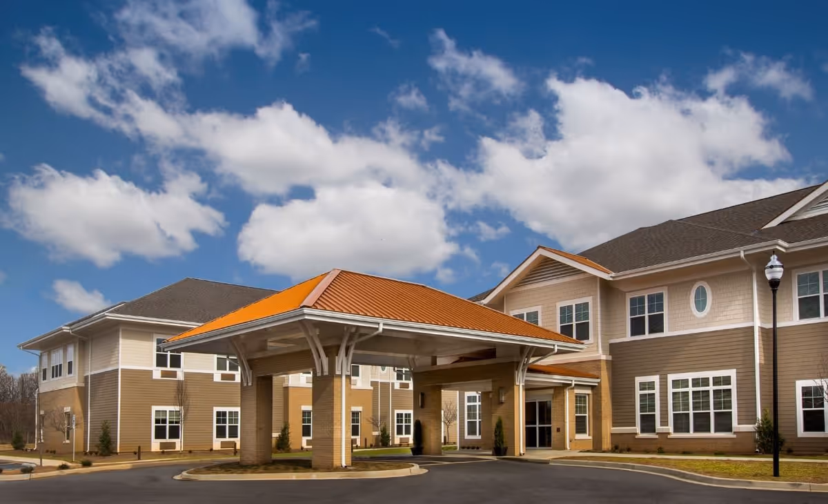 Exterior view of a senior living facility named The Residences at Greer, showing a large covered entrance with an orange roof, beige and brown building walls, multiple windows, and a clear blue sky with scattered clouds.