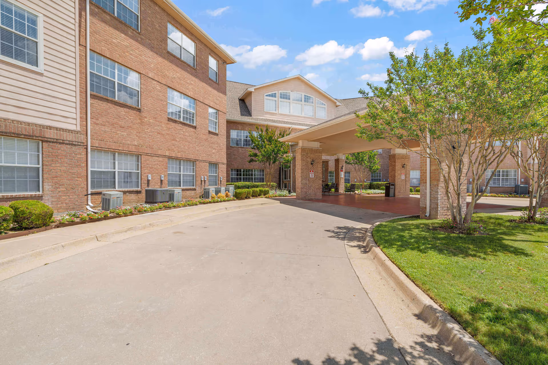 Exterior view of Morada Cedar Hill senior living facility showing a brick building with multiple windows, a covered entrance with pillars, a driveway, and landscaped greenery including trees and bushes under a partly cloudy sky.