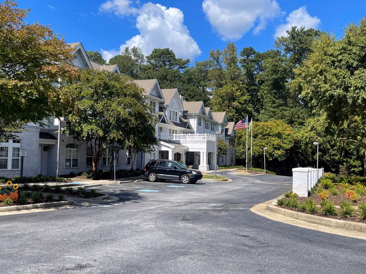 Front exterior of a multi-story senior living building with a driveway, a parked car, an American flag, and trees under a blue sky.