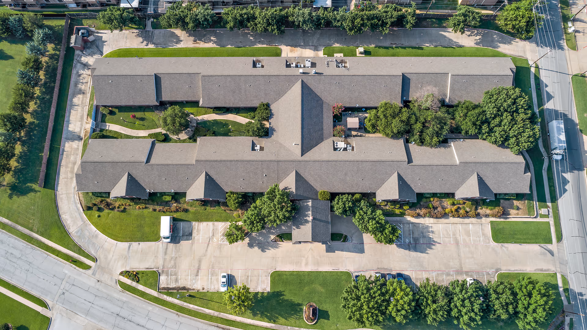 Aerial view of Morada North Richland Hills facility showing a large, single-story building with a gray roof surrounded by green lawns, trees, and parking areas. The building has a central entrance with a covered drop-off area and landscaped pathways within the property.