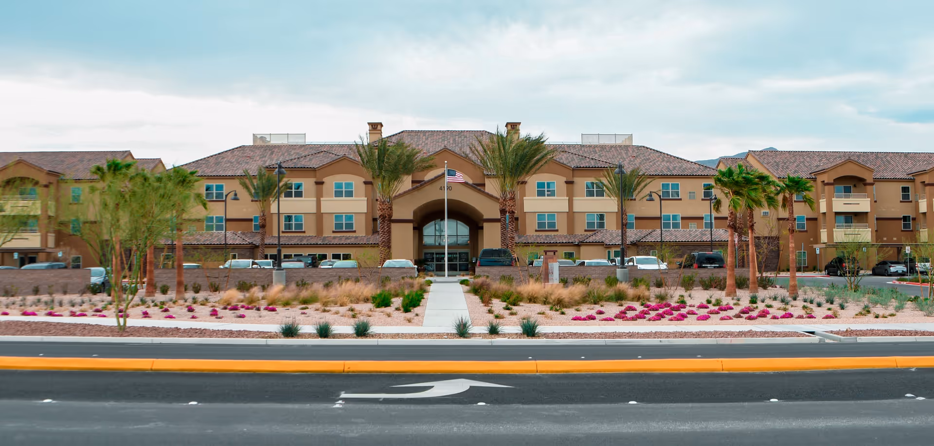 Front exterior view of Vista Park Retirement Community building with a central entrance, palm trees, landscaped garden with flowers and shrubs, and a road with a directional arrow in the foreground.
