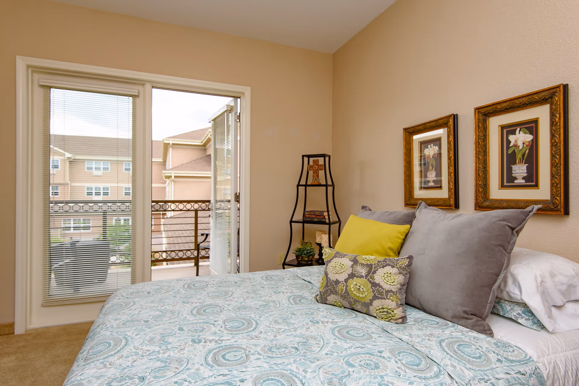 A bedroom with a bed covered in a blue and white patterned bedspread and several pillows in gray, yellow, and floral designs. Two framed floral pictures hang on the beige wall above the bed. A black metal corner shelf holds a cross, books, a small plant, and a lamp. A glass door with blinds opens to a balcony with a view of neighboring buildings.