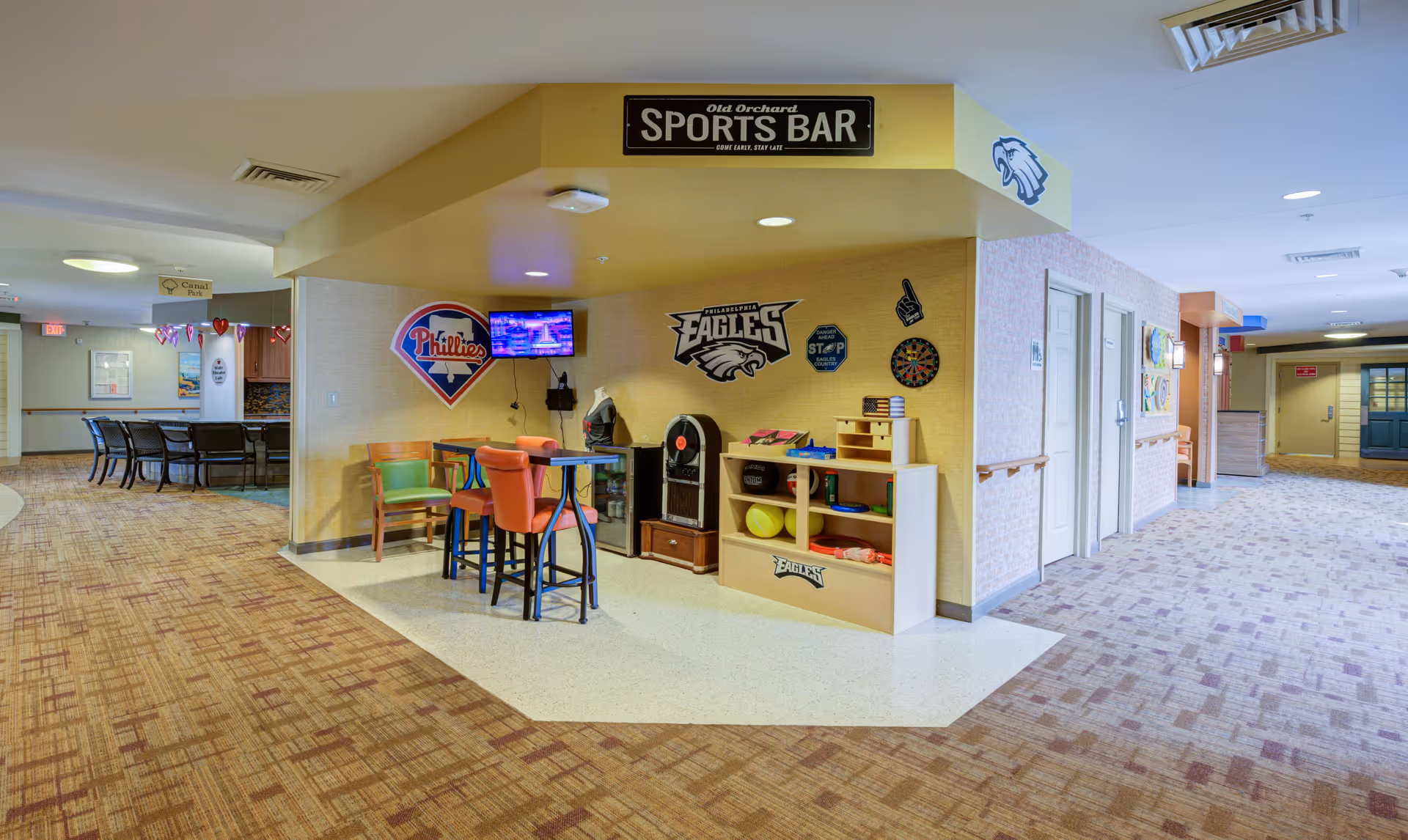 Interior view of a senior living facility hallway featuring a small sports bar area with a high table and chairs, sports memorabilia on the walls including Philadelphia Eagles and Phillies logos, a dartboard, and a TV mounted on the wall. The hallway has carpeted floors, handrails along the walls, and several doors leading to other rooms.
