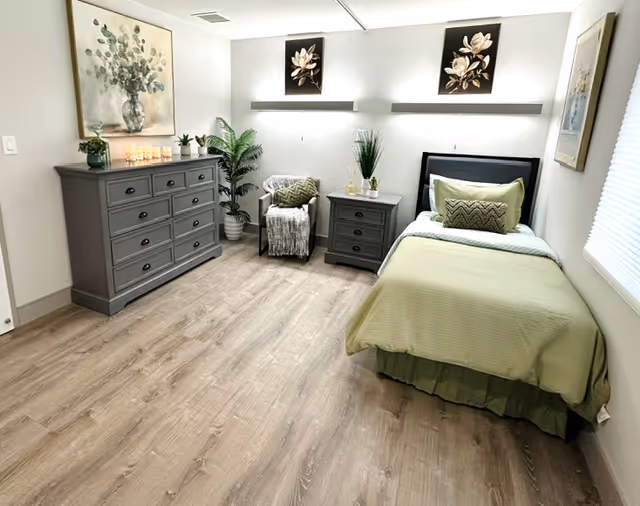 A neatly arranged bedroom with a single bed covered in light green bedding and pillows. Next to the bed is a gray nightstand with decorative plants. Across from the bed is a matching gray dresser with candles and a plant on top. A comfortable chair with a throw blanket is positioned in the corner near a tall potted plant. The walls are decorated with floral artwork and the floor is light wood.