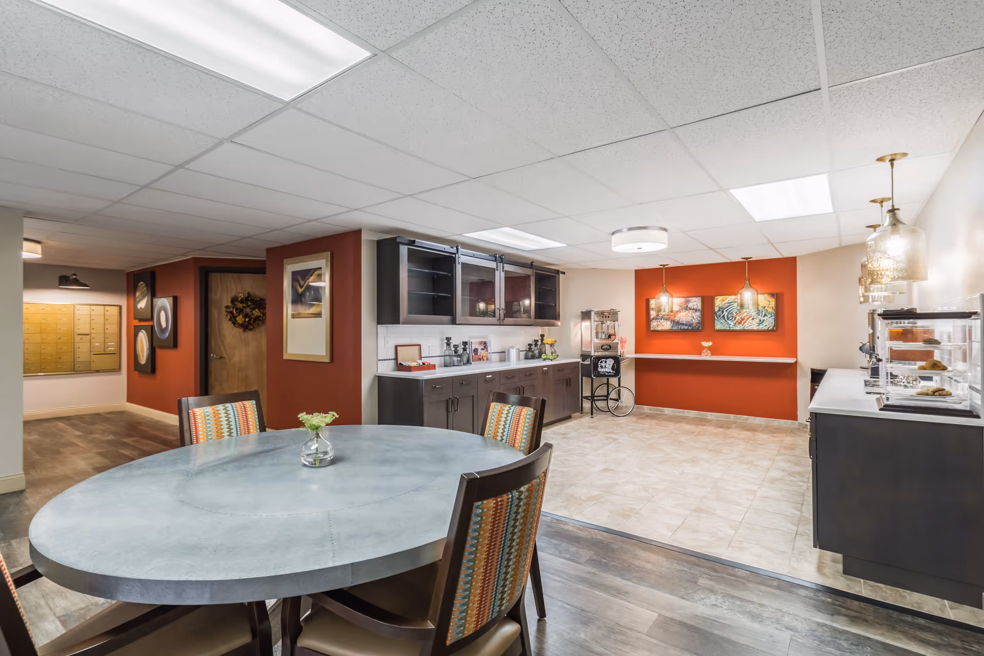 Communal dining and refreshment area with a round table and chairs, kitchenette cabinets, and a service counter against an orange accent wall.