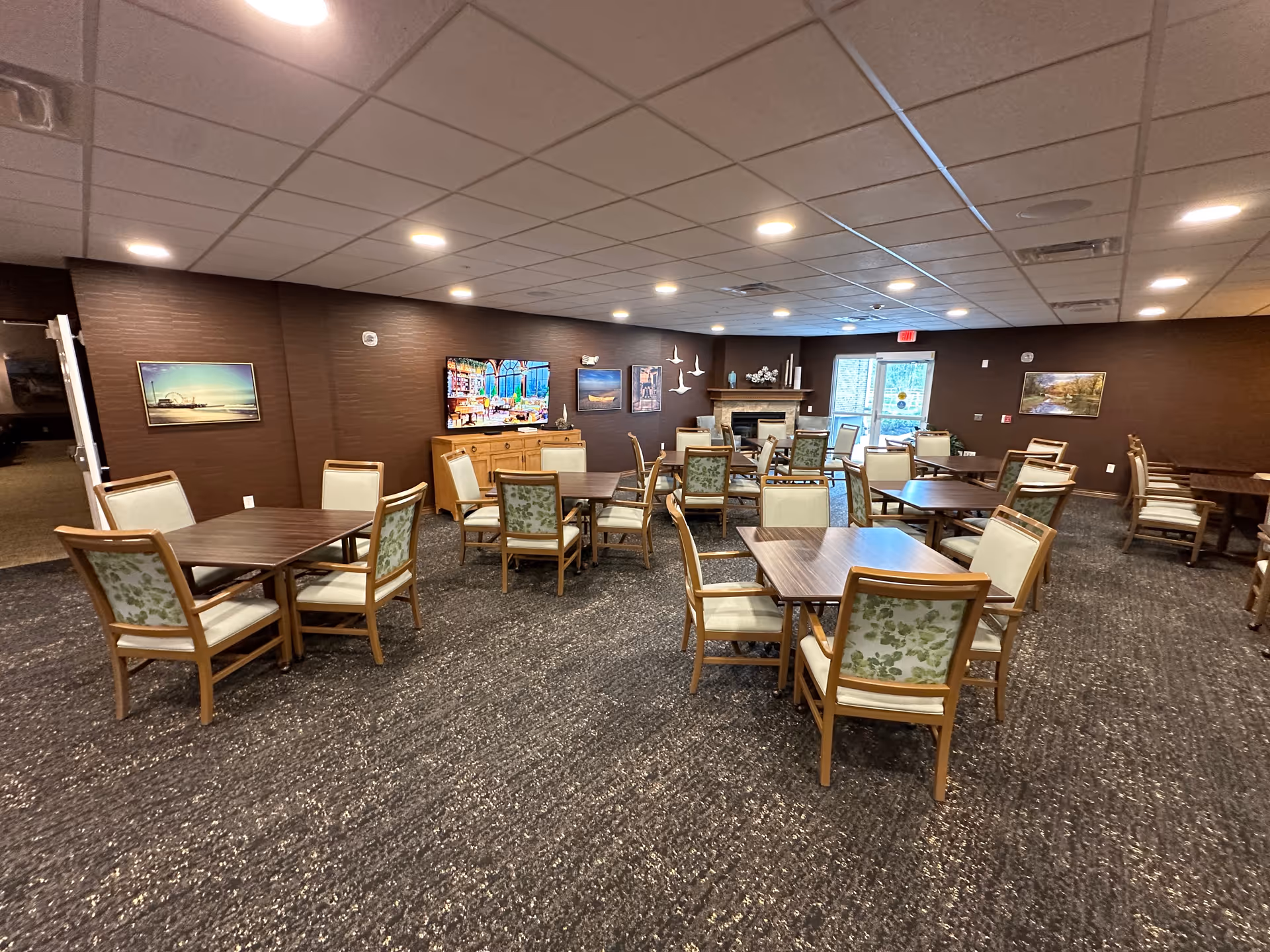 A spacious dining room in a retirement community with multiple wooden tables and chairs arranged neatly. The chairs have cushioned seats and backs with a floral pattern. The room has a carpeted floor, brown walls adorned with framed pictures, a fireplace, and a large window letting in natural light.