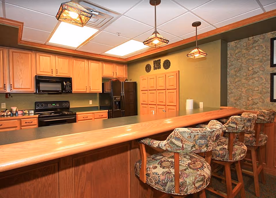 A kitchen-style bar area with wooden cabinets, black appliances, and patterned bar stools along a long countertop.