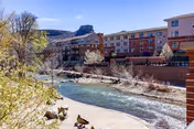 A scenic outdoor view of a river flowing alongside a multi-story residential building with balconies. Trees and shrubs line the riverbank, and a rocky hill or mesa is visible in the background under a clear sky.