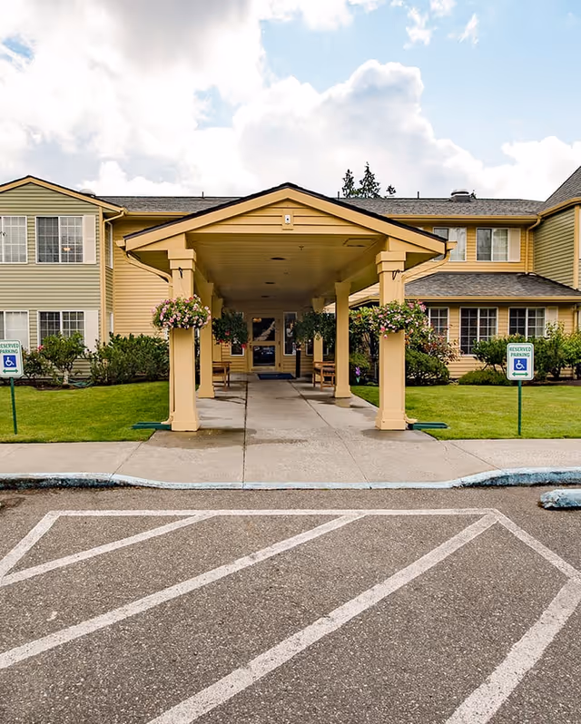 Entrance to a senior living facility with a covered walkway supported by columns adorned with hanging flower baskets. The building is two stories with beige and light green siding, surrounded by well-maintained grass and shrubs. There are reserved parking signs for handicapped parking on either side of the walkway.