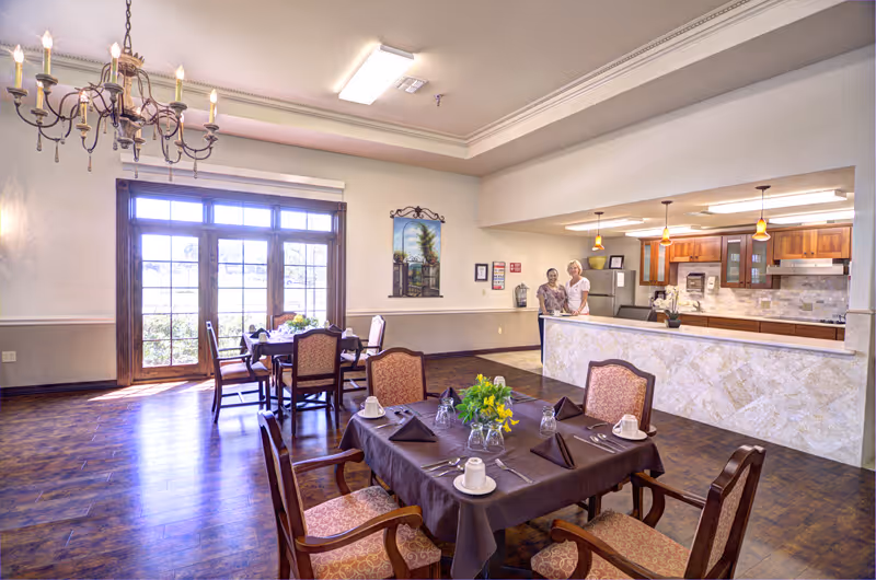 A bright dining room in a senior living facility with wooden floors and large windows letting in natural light. There are two dining tables set with dark tablecloths, napkins, cups, and a centerpiece of yellow flowers. In the background, two women stand behind a kitchen counter with wooden cabinets and pendant lights hanging above.