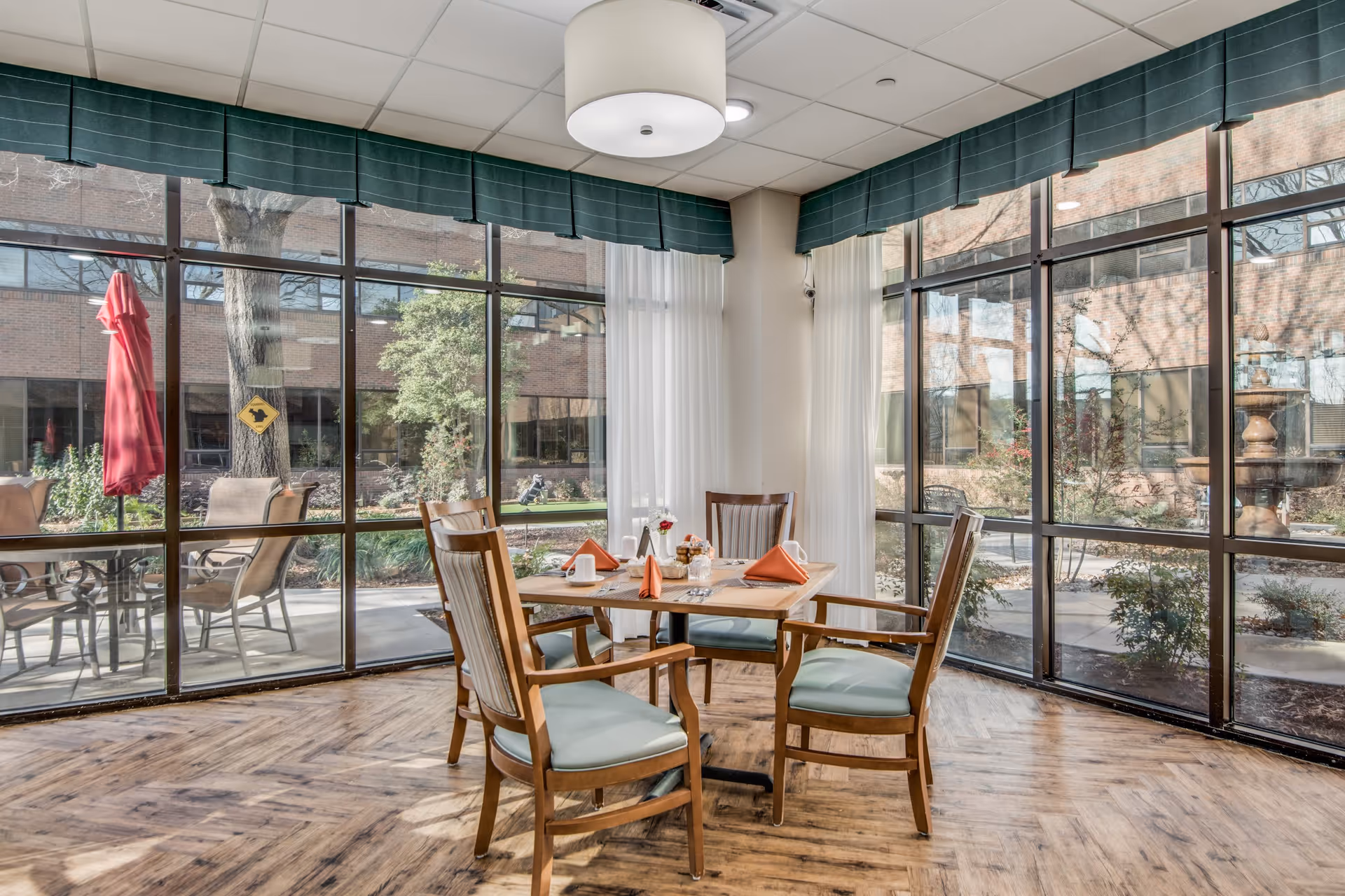 A bright dining area with a square wooden table set for four, featuring orange folded napkins, white cups, and a small flower vase. The room has large floor-to-ceiling windows with green valances and white curtains, offering a view of an outdoor patio with chairs, a red umbrella, and greenery.