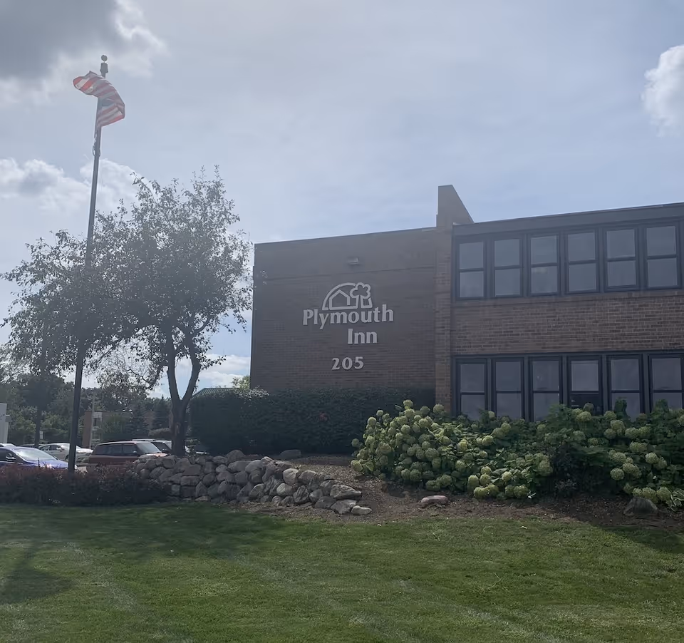 Exterior view of a brick building with the sign 'Plymouth Inn 205' on the wall, surrounded by green bushes and a tree. An American flag is flying on a flagpole to the left, and there are cars parked in the background under a partly cloudy sky.