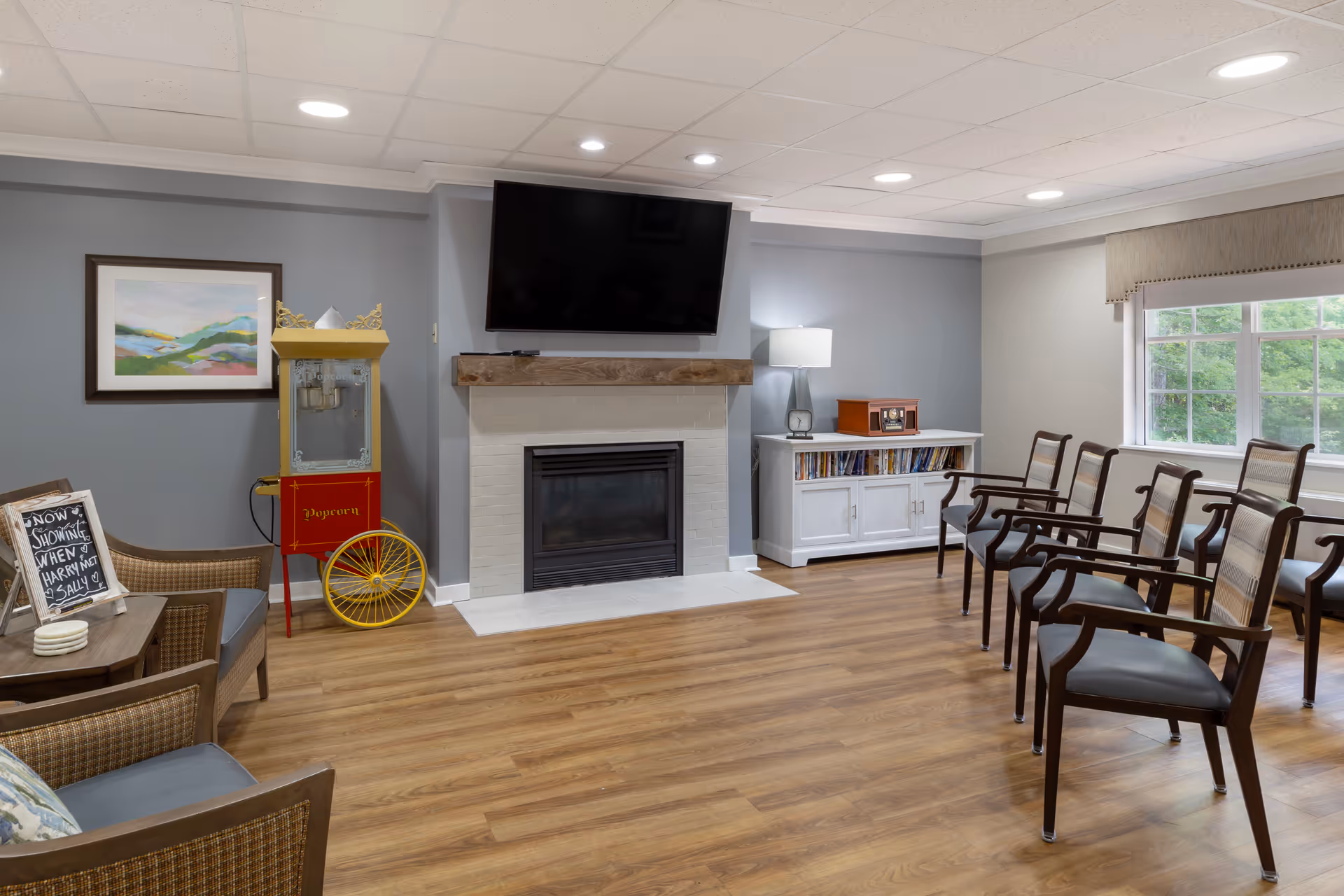 A cozy common area with wooden flooring, a fireplace with a mounted flat-screen TV above it, a vintage popcorn machine, a white cabinet with books and a lamp, several wooden chairs arranged in rows, a window with a view of greenery outside, and a small chalkboard sign on a table.