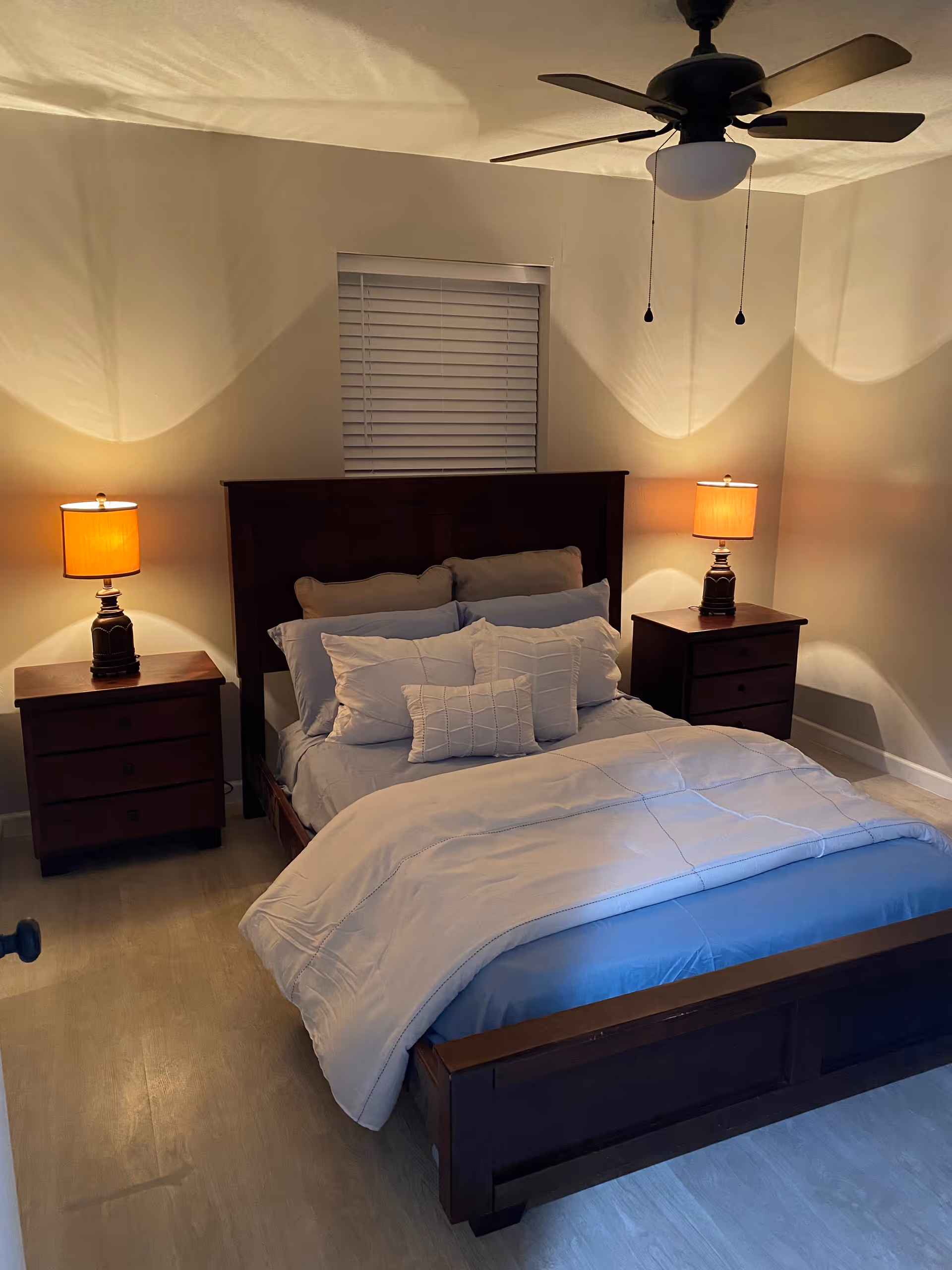 A cozy bedroom with a wooden bed frame and headboard, neatly made with light-colored bedding and multiple pillows. There are two matching wooden nightstands on either side of the bed, each with a lit table lamp. A ceiling fan with a light fixture is mounted on the ceiling, and a window with closed blinds is centered on the wall behind the bed.