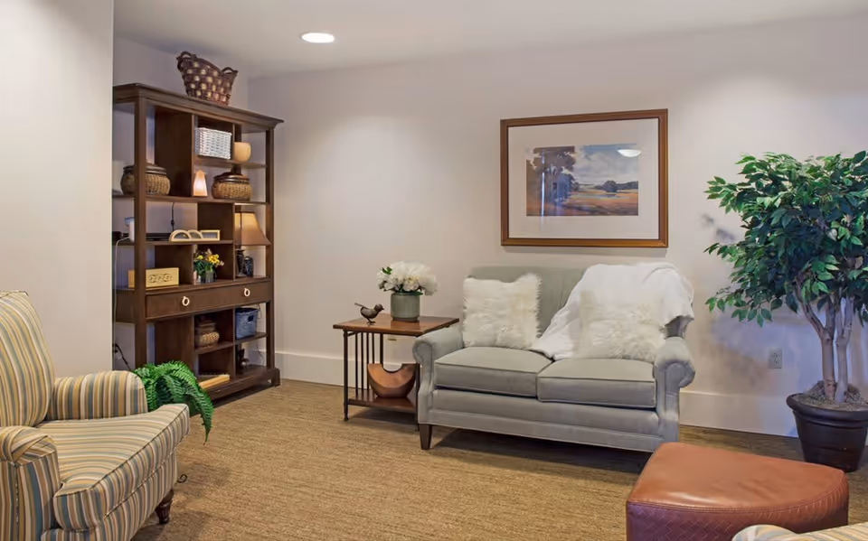 A cozy living room area with a light gray loveseat adorned with two fluffy white pillows and a white throw blanket. Next to the loveseat is a wooden side table with a vase of white flowers and a small bird figurine. To the left, there is a striped armchair and a dark wooden shelving unit filled with decorative baskets, books, and a lamp. A large potted plant is placed on the right side of the loveseat, and a framed landscape painting hangs on the wall above it. The room has beige carpeting and soft lighting.