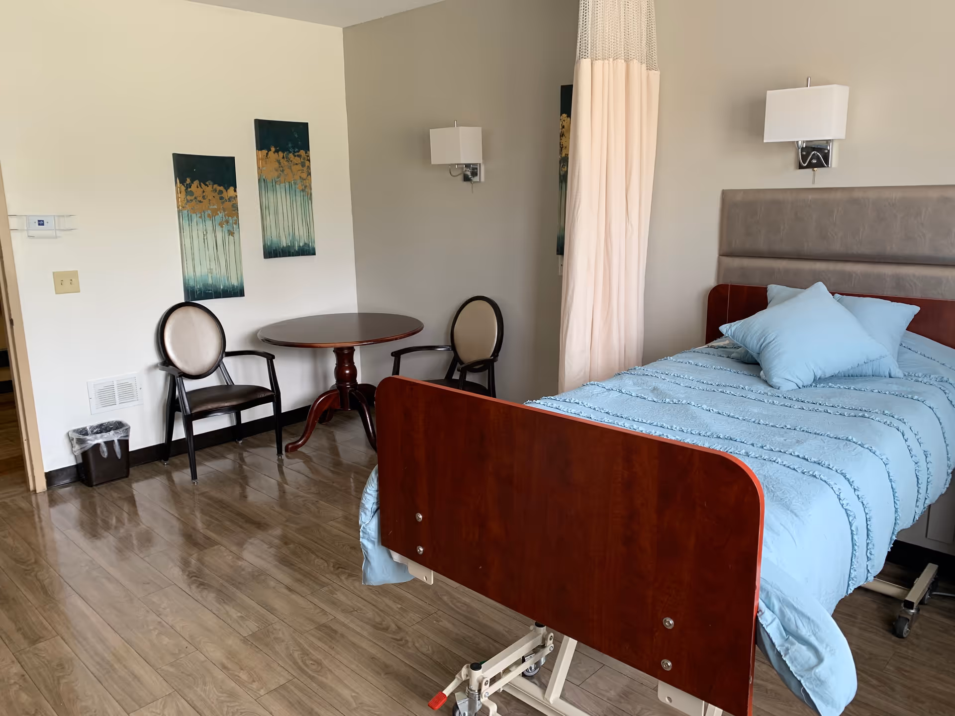 A nursing facility bedroom with a hospital bed covered in a light blue blanket and pillows. Next to the bed is a privacy curtain. On the opposite wall, there is a small round wooden table with two chairs. The floor is wood laminate, and two vertical paintings with abstract designs hang on the wall.